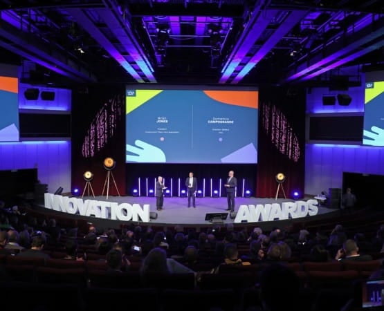 Three people with microphones and a giant screen on a stage with giant letters placed on the stage in front of a crowd at the INTERMAT Innovation Awards 2024